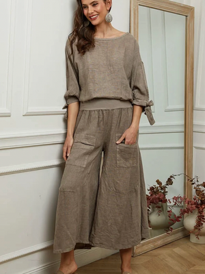 Woman wearing a brown top and wide leg linen pants standing in a room with a mirror and decorative items.