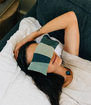 Person lying on a couch with a green striped eye mask on, wearing a white shirt.