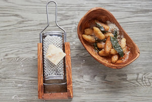 Wood and metal cheese grater with a block of cheese next to a wooden bowl of food on a wooden surface