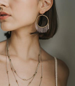 Close-up of a person wearing gold hoop earrings and necklaces against a neutral background
