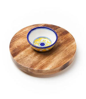 Wooden round cheese board with a ceramic bowl featuring blue and yellow patterns on a white background