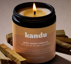 Kandu Palo Santo Tobacco candle in a jar with a lit wick, surrounded by wooden sticks on a beige background.
