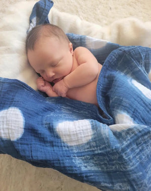 Newborn baby wrapped in a blue tie-dye swaddle on a light surface.