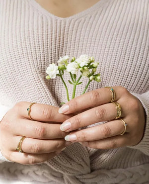 Close-up of hands holding a small plant with white flowers, wearing gold stacking rings.