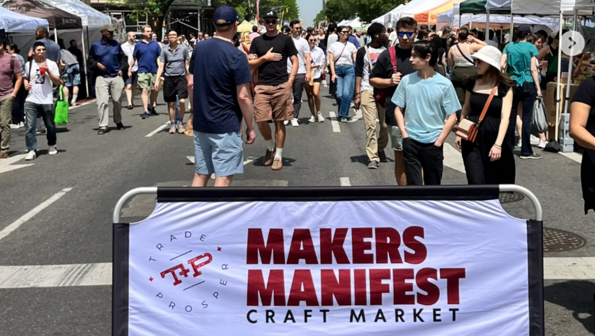 People walking on a street with a 'Makers Manifest Craft Market' sign in the foreground.