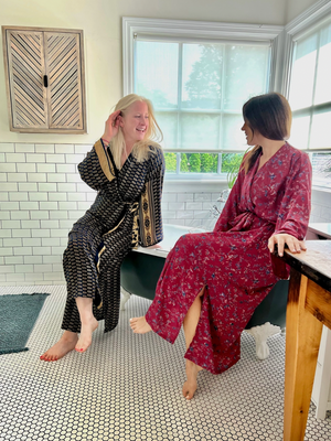Two women in patterned robes sitting on a claawfoot tub in a bathroom with large windows.