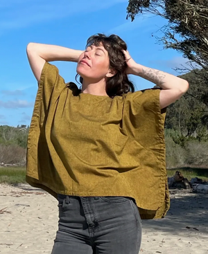 Woman wearing a mustard yellow boxy top and dark jeans standing on a beach with trees and blue sky in the background.
