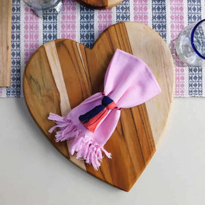 Heart-shaped wooden cutting board with a pink napkin on top on a patterned tablecloth.