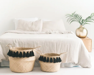 Two woven baskets with black tassels on a white surface in front of a bed.