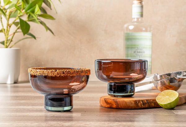 Two brown glasses on a wooden surface with a bottle and lime in the background.