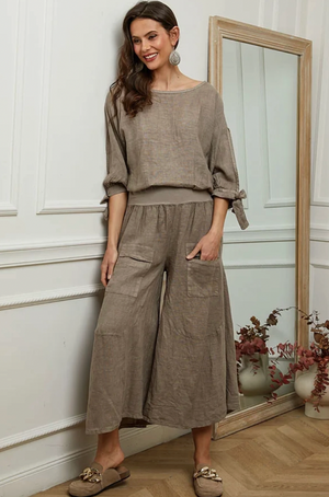 Woman wearing a brown top and wide leg linen pants standing in a room with a mirror and decorative items.