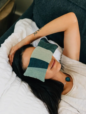 Person lying on a couch with a green striped eye mask on, wearing a white shirt.