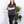 Woman wearing a dark gray henley shirt and black pants standing in a room with white walls and a plant.