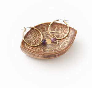 Gold hoop earrings with purple beads on a wooden dish with eye design