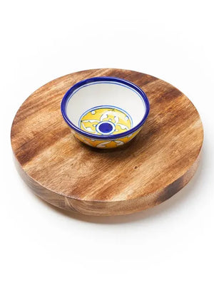 Wooden round cheese board with a ceramic bowl featuring blue and yellow patterns on a white background