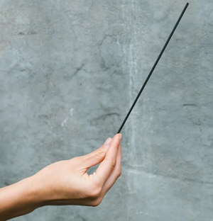 Hand holding a black incense stick against a gray concrete wall