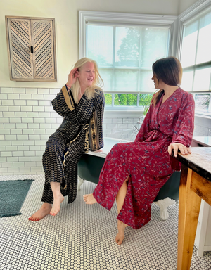 Two women in patterned robes sitting on a claawfoot tub in a bathroom with large windows.