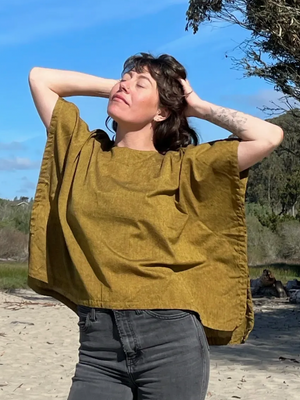 Woman wearing a mustard yellow boxy top and dark jeans standing on a beach with trees and blue sky in the background.