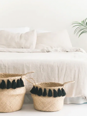 Two woven baskets with black tassels on a white surface in front of a bed.